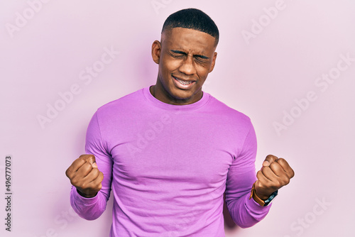 Young black man wearing casual pink sweater very happy and excited doing winner gesture with arms raised, smiling and screaming for success. celebration concept.