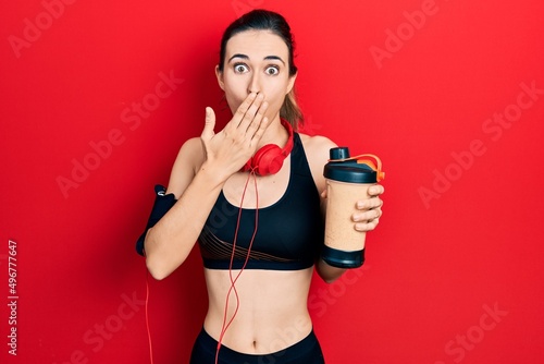 Young hispanic girl wearing sport clothes drinking a protein shake covering mouth with hand, shocked and afraid for mistake. surprised expression