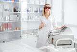 Cheerful young woman beautician standing near laser epilation machine