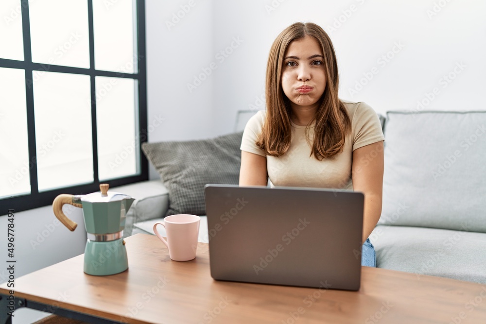 Young brunette woman using laptop at home drinking a cup of coffee puffing cheeks with funny face. mouth inflated with air, crazy expression. Young brunette woman using laptop at home drinking a cup of coffee puffing cheeks with funny face. mouth inflated with air, crazy expression.