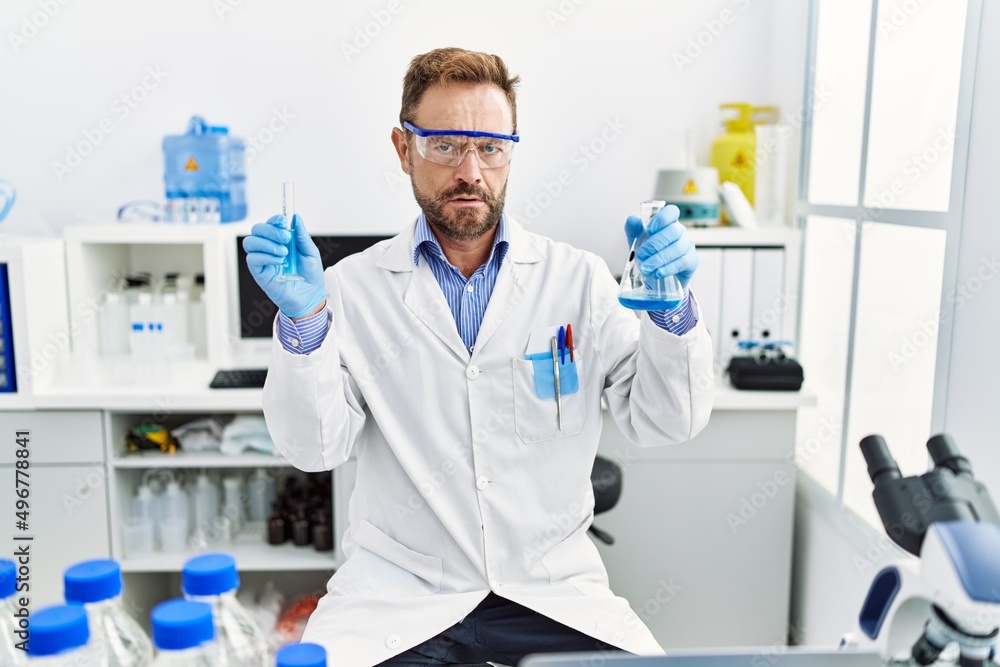 Middle age man working at scientist laboratory holding chemical products clueless and confused expression. doubt concept. Middle age man working at scientist laboratory holding chemical products clueless and confused expression. doubt concept.