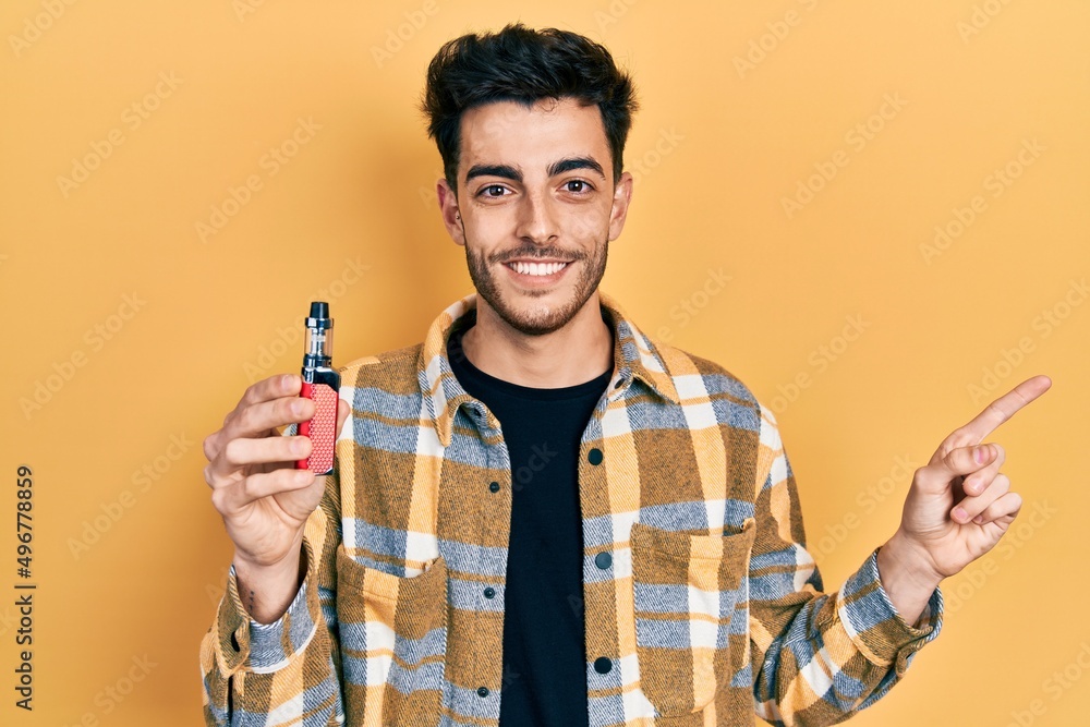 Young hispanic man holding electronic cigarette smiling happy pointing with hand and finger to the side Young hispanic man holding electronic cigarette smiling happy pointing with hand and finger to the side