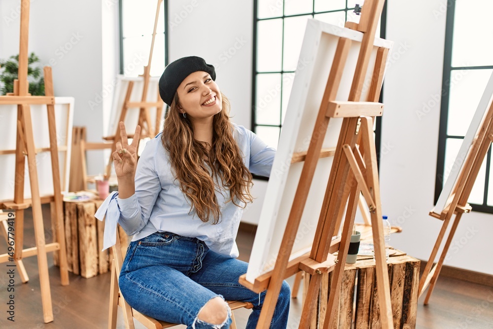 Young hispanic artist woman painting on canvas at art studio smiling looking to the camera showing fingers doing victory sign. number two. Young hispanic artist woman painting on canvas at art studio smiling looking to the camera showing fingers doing victory sign. number two.