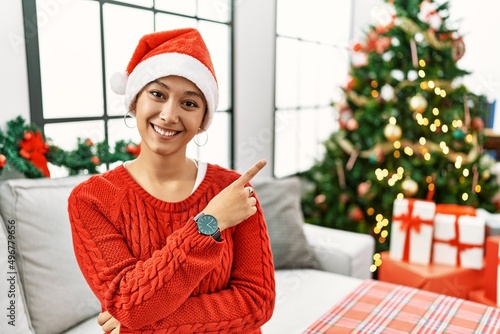 Young hispanic woman with short hair wearing christmas hat sitting on the sofa with a big smile on face, pointing with hand finger to the side looking at the camera.