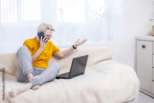 adult woman is emotionally talking on the phone while sitting at a laptop on the couch at home © Irina