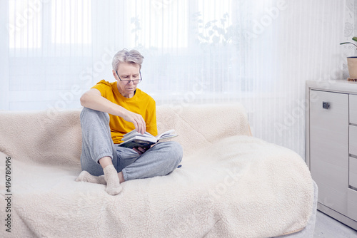  adult gray-haired woman with glasses in home clothes reads a book sitting on the couch at home