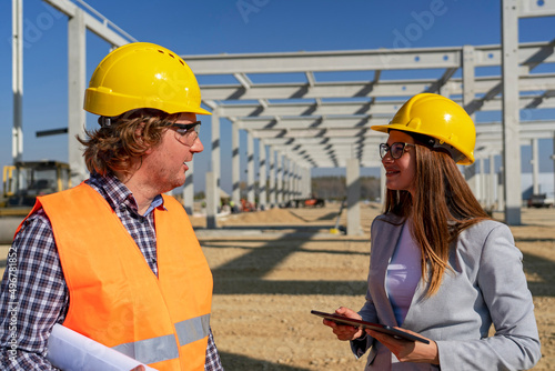 Young Female Architect Using Digital Tablet And Having Conversation With Construction Foreman At Construction Site. Man and Woman in Yellow Hardhats Discuss a Construction Project. Gender Equality. 