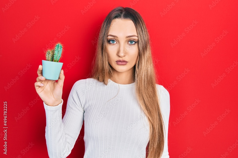 Young blonde girl holding small cactus pot thinking attitude and sober expression looking self confident Young blonde girl holding small cactus pot thinking attitude and sober expression looking self confident