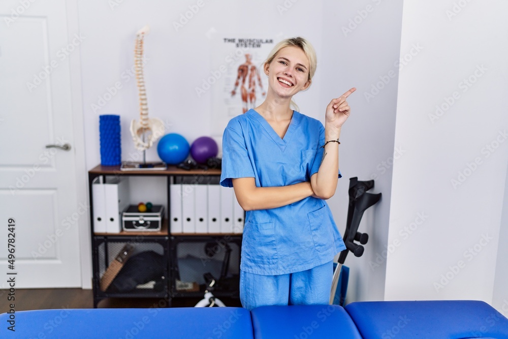 Young physiotherapist woman working at pain recovery clinic with a big smile on face, pointing with hand finger to the side looking at the camera. Young physiotherapist woman working at pain recovery clinic with a big smile on face, pointing with hand finger to the side looking at the camera.