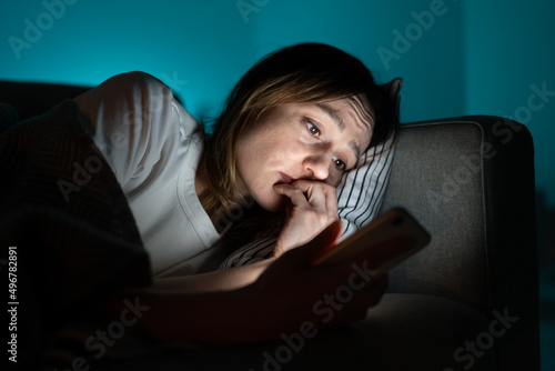 Young anxious woman lying in bed staring at smartphone screen at night, reading about depression symptoms in internet, phone addicted female can not stop scrolling news media before bedtime. Anxiety.