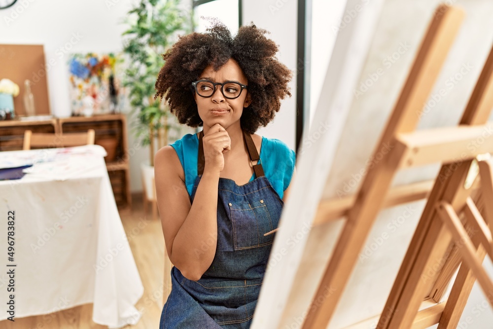 Beautiful african american woman with afro hair painting canvas at art studio serious face thinking about question with hand on chin, thoughtful about confusing idea Beautiful african american woman with afro hair painting canvas at art studio serious face thinking about question with hand on chin, thoughtful about confusing idea