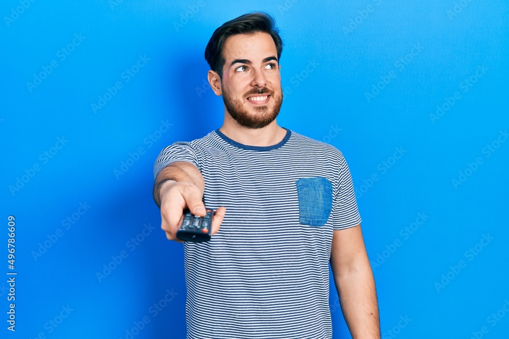 Handsome caucasian man with beard holding television remote control smiling looking to the side and staring away thinking. Handsome caucasian man with beard holding television remote control smiling looking to the side and staring away thinking.