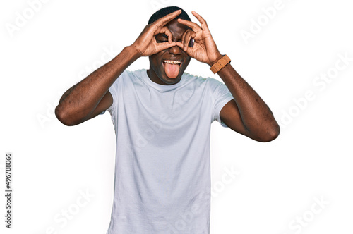 Young african american man wearing casual white t shirt doing ok gesture like binoculars sticking tongue out, eyes looking through fingers. crazy expression.