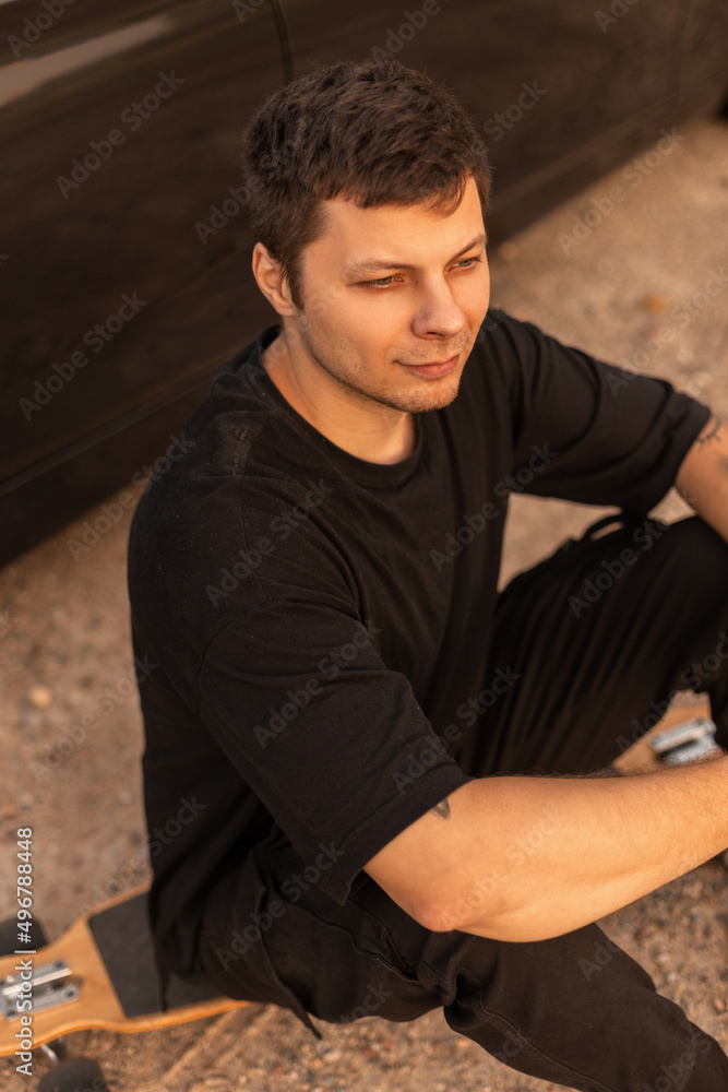 Hipster man in black t-shirt and black pants sits and relax on longboard outdoors. Hipster man in black t-shirt and black pants sits and relax on longboard outdoors.