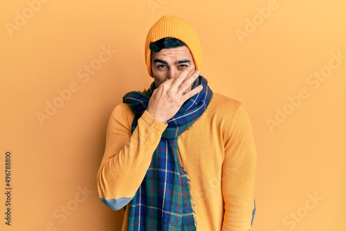 Handsome hispanic man wearing wool hat and winter scarf smelling something stinky and disgusting, intolerable smell, holding breath with fingers on nose. bad smell © Krakenimages.com