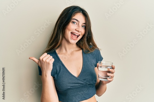 Canvas Print Young caucasian woman drinking glass of water pointing thumb up to the side smil