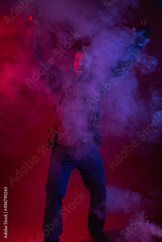 soldier in full gear with weapons. a man in headphones, body armor, with a backpack and a belt. red background. colored, blue-red light. smoke around the military. explosion, chemical attack