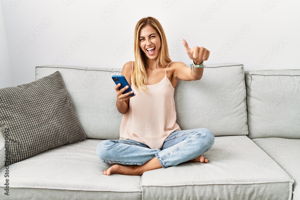 Blonde beautiful young woman sitting on the sofa at home using smartphone approving doing positive gesture with hand, thumbs up smiling and happy for success. winner gesture.