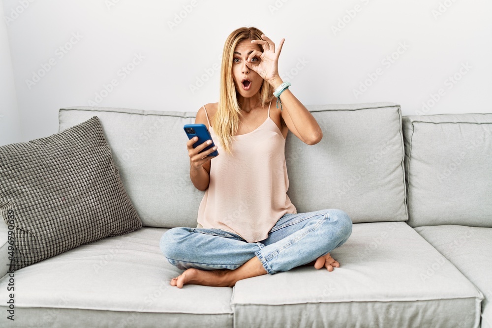 Blonde beautiful young woman sitting on the sofa at home using smartphone doing ok gesture shocked with surprised face, eye looking through fingers. unbelieving expression. Blonde beautiful young woman sitting on the sofa at home using smartphone doing ok gesture shocked with surprised face, eye looking through fingers. unbelieving expression.