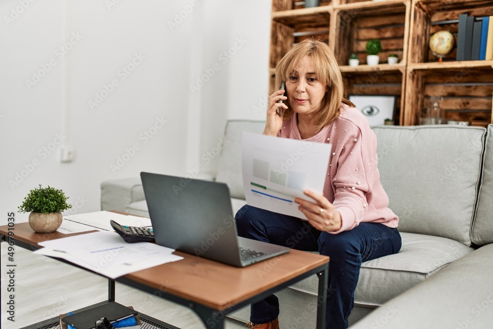 Fototapeta premium Middle age blonde woman teleworking using laptop and talking on the smartphone at home.