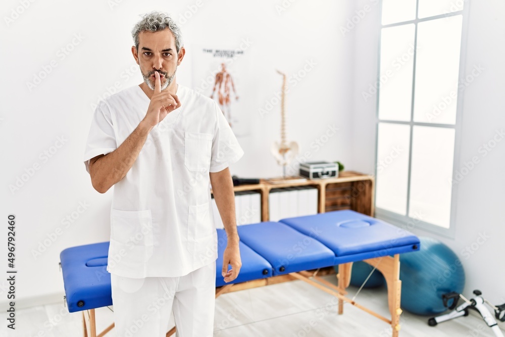 Middle age hispanic therapist man working at pain recovery clinic asking to be quiet with finger on lips. silence and secret concept. Middle age hispanic therapist man working at pain recovery clinic asking to be quiet with finger on lips. silence and secret concept.