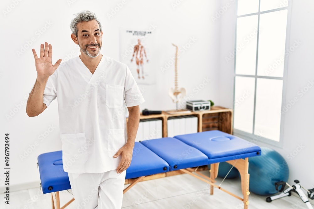 Middle age hispanic therapist man working at pain recovery clinic waiving saying hello happy and smiling, friendly welcome gesture Middle age hispanic therapist man working at pain recovery clinic waiving saying hello happy and smiling, friendly welcome gesture