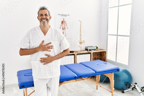 Middle age hispanic therapist man working at pain recovery clinic smiling and laughing hard out loud because funny crazy joke with hands on body.