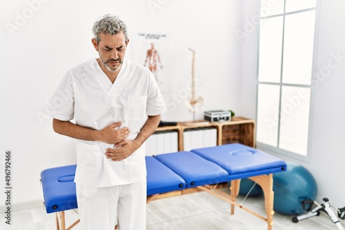 Middle age hispanic therapist man working at pain recovery clinic with hand on stomach because indigestion  painful illness feeling unwell. ache concept.