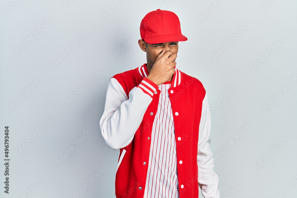 Young african american man wearing baseball uniform smelling something stinky and disgusting, intolerable smell, holding breath with fingers on nose. bad smell Young african american man wearing baseball uniform smelling something stinky and disgusting, intolerable smell, holding breath with fingers on nose. bad smell