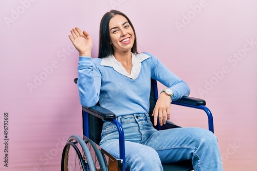 Beautiful woman with blue eyes sitting on wheelchair waiving saying hello happy and smiling, friendly welcome gesture