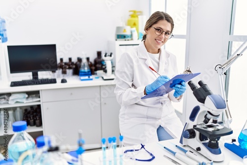 Young hispanic woman wearing scientist uniform using microscope at laboratory