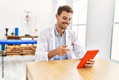 Young hispanic man wearing physiotherapist uniform using touchpad at clinic