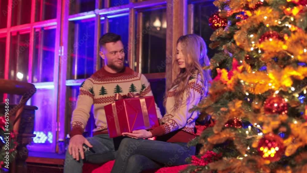 Young beautiful couple sitting at the windowsill of a country house near Christmas tree. Girl is holding a gift box and shaking it to guess what's inside. Young beautiful couple sitting at the windowsill of a country house near Christmas tree. Girl is holding a gift box and shaking it to guess what's inside.