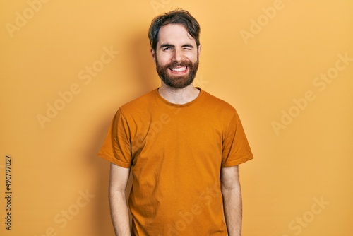 Caucasian man with beard wearing casual yellow t shirt winking looking at the camera with sexy expression, cheerful and happy face.