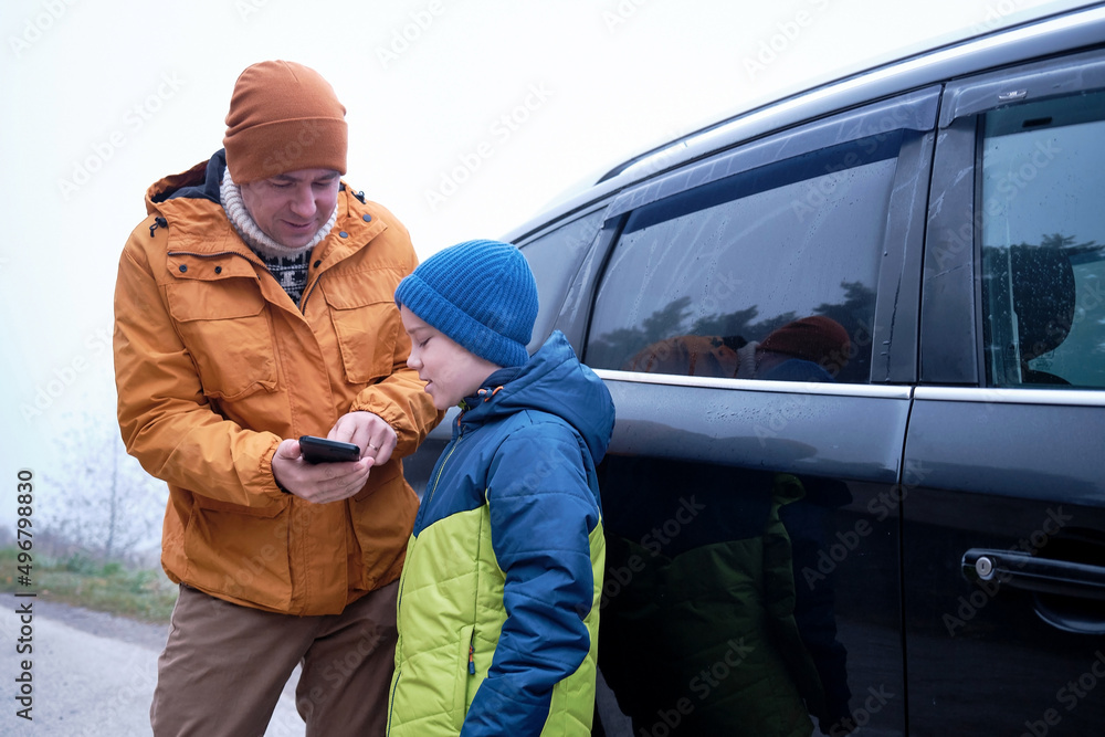 Happy father and son standing together near new car on a journey. Family are resting on the side of the road on a road trip. Child takes pictures on smartphone. Happy family travels.