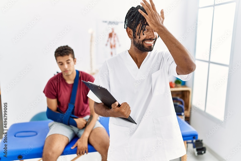 Young hispanic man working at pain recovery clinic with a man with broken arm surprised with hand on head for mistake, remember error. forgot, bad memory concept. Young hispanic man working at pain recovery clinic with a man with broken arm surprised with hand on head for mistake, remember error. forgot, bad memory concept.