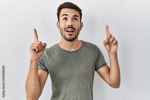 Young hispanic man with beard wearing casual t shirt over white background amazed and surprised looking up and pointing with fingers and raised arms.