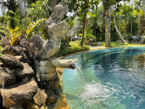 Old statue shaped fountain covered with moss and dry leaves, small pond in a tropical park. Clear blue water. Landscape design element. South garden. Antique sculpture. Green trees, foliage, grass.