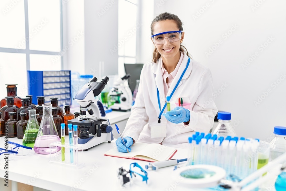 Fototapeta premium Young hispanic woman wearing scientist uniform writing on notebook and holding test tube at laboratory