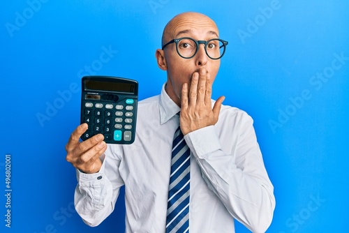 Bald business man with beard showing calculator device covering mouth with hand, shocked and afraid for mistake. surprised expression