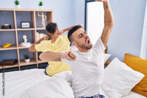 Two man couple waking up stretching arms sitting on bed at bedroom