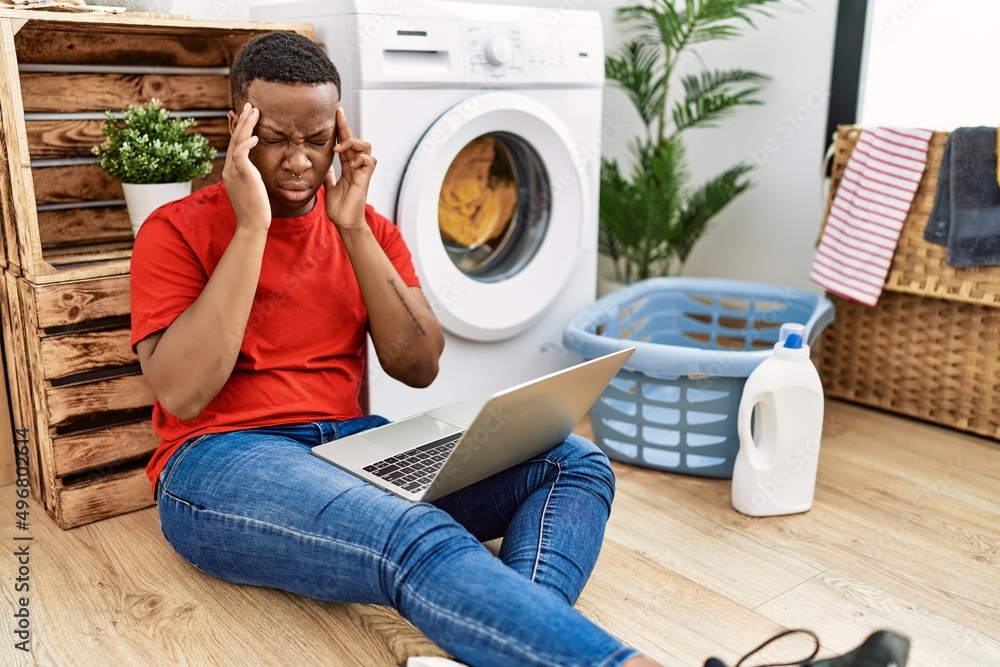 Young african man doing laundry and using computer with hand on head for pain in head because stress. suffering migraine. Young african man doing laundry and using computer with hand on head for pain in head because stress. suffering migraine.