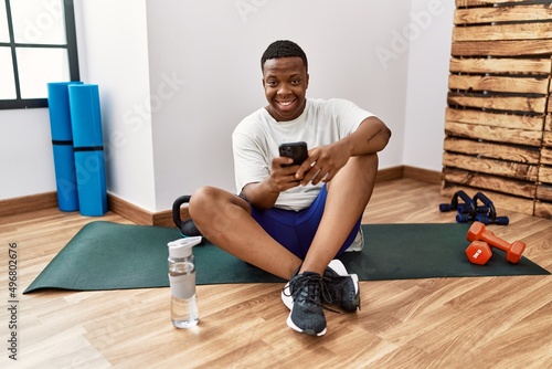 Young african man sitting on training mat at the gym using smartphone with a happy and cool smile on face. lucky person.