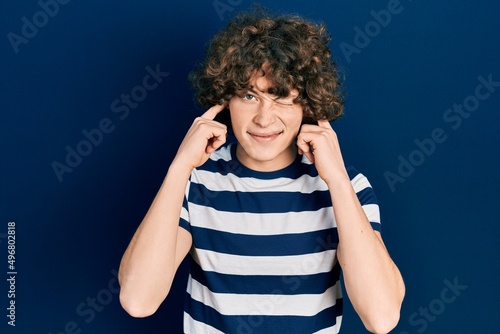 Handsome young man wearing casual striped t shirt covering ears with fingers with annoyed expression for the noise of loud music. deaf concept.