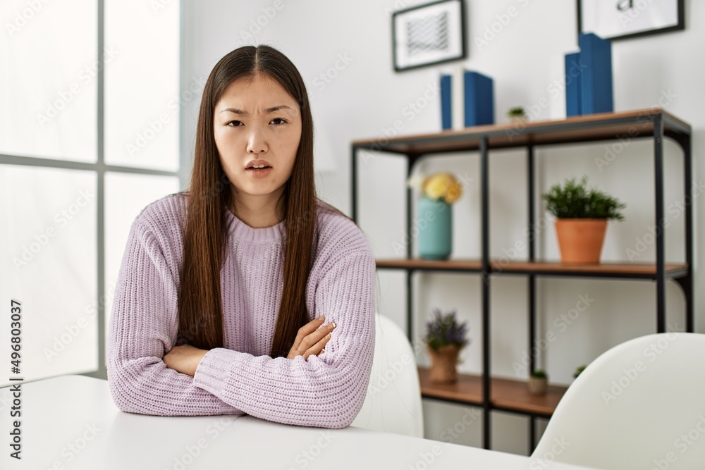 Young chinese girl wearing casual clothes sitting on the table at home skeptic and nervous, disapproving expression on face with crossed arms. negative person.