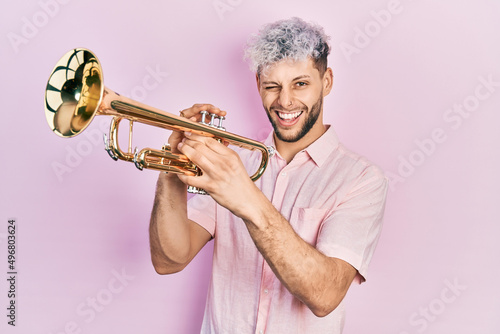 Canvas Print Young hispanic man with modern dyed hair playing trumpet winking looking at the camera with sexy expression, cheerful and happy face