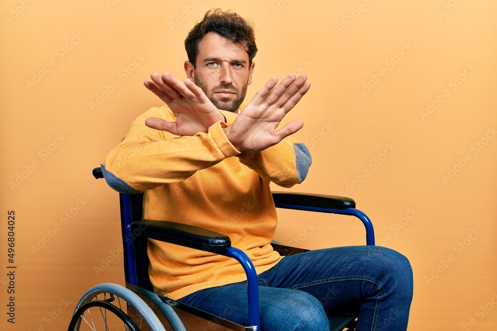 Handsome man with beard sitting on wheelchair rejection expression crossing arms doing negative sign, angry face