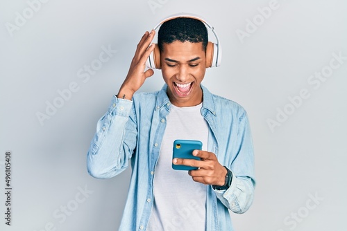 Young african american guy using smartphone wearing headphones celebrating victory with happy smile and winner expression with raised hands