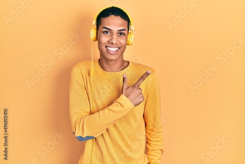 Young african american guy listening to music using headphones cheerful with a smile on face pointing with hand and finger up to the side with happy and natural expression