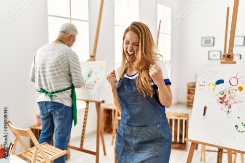 Hispanic woman wearing apron at art studio very happy and excited doing winner gesture with arms raised, smiling and screaming for success. celebration concept.